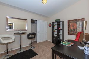 A cozy room featuring a small desk with a black chair, a set of two modern bar stools, and a round table. The walls are painted beige with a mirror and artwork displayed. A bookshelf with decorative items stands against one wall, and a closet is visible through an open door. Soft lighting creates a warm atmosphere.
