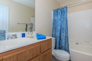 A bright bathroom featuring a white bathtub with a blue shower curtain, a wooden vanity with a sink, and decorative items including a seashell and a seahorse statue. Blue towels are neatly arranged on the countertop, and there is a mirror reflecting the space. The overall decor has a coastal theme.