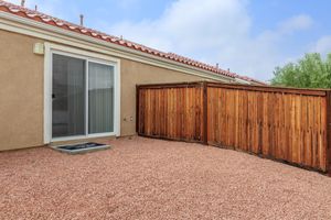 A small outdoor area featuring a sliding glass door, bordered by wooden fences. The ground is covered with small gravel stones, and a welcome mat is placed at the entrance. The space is framed by walls with a tiled roof and is surrounded by greenery in the background.