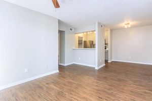 Spacious, well-lit living area with a ceiling fan, featuring light-colored walls and hardwood-style flooring. An open layout connects to a modern kitchen with stainless steel appliances. Natural light streams in from nearby windows, creating a welcoming atmosphere.