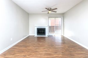 A spacious and empty living room featuring light-colored walls, a ceiling fan, a white fireplace, and large sliding glass doors leading to an outdoor space. The floor is made of wood, and the lighting appears bright and inviting. There are no furnishings present.