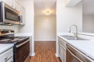 View of a modern kitchen featuring stainless steel appliances, including a microwave and oven, white countertops, and a sink. The kitchen has wood-style flooring and leads to an adjacent room with neutral-colored walls and a ceiling light.