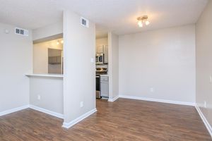 An interior view of a modern apartment with hardwood-style flooring. The space includes an open layout connecting the kitchen to a living area. The kitchen features grey cabinets and appliances, while the living area is bright with natural light and a ceiling fixture. The walls are painted in soft neutral tones.