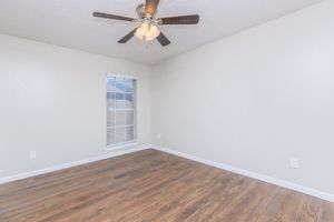 A spacious, empty room with light-colored walls and brown wooden flooring. A ceiling fan with light fixtures hangs from the ceiling. There is a window with blinds on one wall, allowing natural light to enter, creating a bright and airy atmosphere.