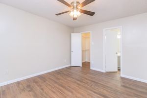 A spacious, empty room featuring light-colored walls and a ceiling fan. The floor is covered with wooden plank flooring. There is an open white door leading to a closet, and another door in the background indicates access to a bathroom. The room has natural light and a clean, neutral aesthetic.