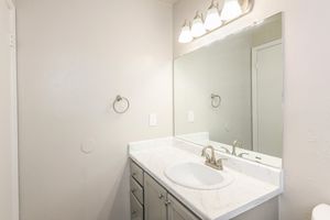 A clean and modern bathroom featuring a white countertop with a sink, a large mirror above, and three light fixtures. The walls are painted a soft gray, and there is a towel holder beside the sink. The cabinetry is a light gray color, adding to the contemporary feel of the space.