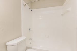 A clean, modern bathroom featuring a white bathtub with a shower, a chrome showerhead, and a white toilet. The walls are tiled in white, creating a bright and airy atmosphere. The space is well-lit and uncluttered, emphasizing a minimalist design.