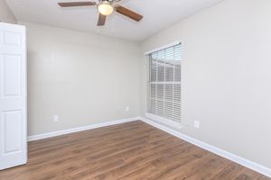 A bright and empty room featuring light-colored walls, a wooden floor, and a ceiling fan. There is a window with blinds providing natural light, and a closet door is visible on the left side of the image. The overall ambiance is calm and inviting, suitable for various uses.