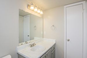A clean and modern bathroom featuring a white countertop with a sink, a large mirror above, and four light fixtures. The walls are painted in a light neutral color, and a door is visible on the right, leading to a separate area. A towel ring hangs next to the sink, and the space appears bright and well-maintained.