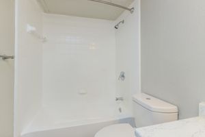 A clean and modern bathroom featuring a white tiled bathtub with a showerhead, a white toilet, and light gray walls. The space is well-lit and minimalistic, emphasizing a fresh and tidy appearance.