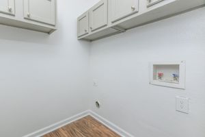A clean, empty laundry room with light gray cabinets mounted on the wall. The wall is painted white, and there are plumbing connections visible, including hot and cold water valves. The floor features light wood-like laminate, contributing to a modern and tidy appearance.
