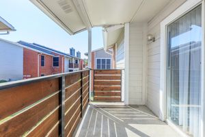 A balcony with a wooden railing, partially covered by a roof. The view shows several neighboring buildings, predominantly in brick and siding materials, under a clear blue sky. Sunlight casts shadows on the floor, creating a bright and inviting atmosphere.