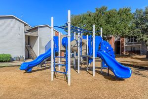 Colorful playground equipment with blue slides and climbing structure on a sandy ground, surrounded by residential buildings and trees under a clear blue sky.