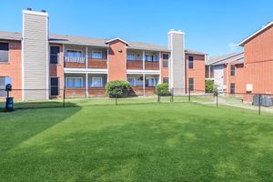 A well-maintained apartment complex featuring red brick buildings with balconies, surrounded by a green lawn. The scene includes a fenced area, clear blue sky, and well-kept landscaping, creating a welcoming atmosphere for residents.