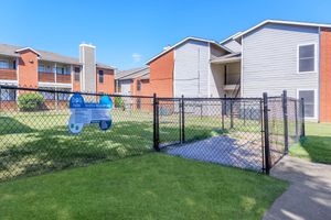 Fenced area labeled "Dog Park" with signage outlining rules and regulations, surrounded by grassy space and residential buildings in the background under a clear blue sky.