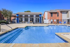 A clear blue swimming pool with a surrounding deck and lounge chairs, adjacent to a two-story building with large windows. The scene is set in a sunny outdoor area with trees in the background and a fence enclosing the pool space.