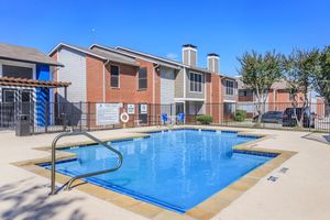 A clean swimming pool surrounded by a paved area, located in front of a two-story brick apartment building. The pool features a metal ladder and is enclosed by a black fence. Visible amenities include lounge chairs and a safety buoy. The sky is clear and blue, indicating a sunny day.