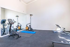 A spacious gym area featuring exercise equipment including an elliptical machine, stationary bike, and weights. There is a blue workout mat on the floor, a mirror reflecting the space, and a generally bright and open atmosphere with light-colored walls.