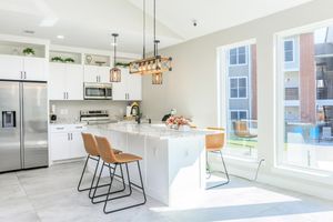 Bright modern kitchen featuring white cabinets, stainless steel appliances, and a large island with seating. A stylish chandelier hangs above the island, and large windows provide natural light, revealing an outdoor view. The space is clean and contemporary, with a neutral color palette.