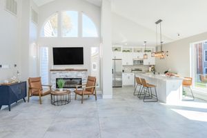 A modern, bright living space featuring a large window with natural light, a sleek television mounted on the wall, a cozy fireplace, and stylish seating. The kitchen area includes a central island with bar stools, emphasizing an open-concept design. Neutral colors and contemporary decor enhance the inviting atmosphere.