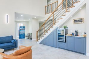Modern interior of a spacious entryway featuring a wooden staircase with metal railings. On one side, a blue couch and a leather chair are arranged around a table. A refreshment area with a mini fridge and snacks is visible, along with a sign that reads “Coffee & Bar.” Bright, natural light illuminates the space.