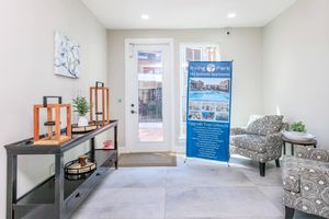 A modern entryway featuring a small table with decorative items, a patterned armchair, and a banner advertising Irving Park apartments. Natural light enters through a large glass door, illuminating the neutral-colored walls and tile flooring.