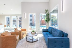A modern lounge space featuring two brown leather armchairs and a blue fabric sofa. A round coffee table sits in the center, surrounded by light-colored walls and large windows that overlook an outdoor pool area. Natural light fills the room, creating a bright and inviting atmosphere.