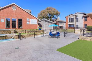 View of an outdoor area featuring a modern swimming pool, lounge chairs, and an umbrella. Surrounding buildings include brick and wood-sided apartments with large windows. Green grass and landscaped areas add to the inviting atmosphere on a sunny day.
