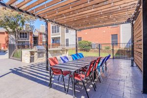 A covered outdoor patio area featuring a large table with red and blue chairs, surrounded by a fence. In the background, residential buildings and a grassy area are visible. Strings of lights hang from the wooden pergola, providing a cozy ambiance for gatherings. A grill is located at one end of the patio.