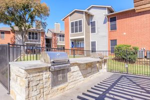 Outdoor grilling area featuring two metal grills on a stone countertop, surrounded by a fenced patio. In the background, there are two residential buildings with a mix of brick and siding, green lawns, and trees, creating a welcoming communal space.