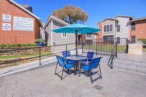 A patio area featuring a round table with four blue chairs under a large turquoise umbrella. In the background, several residential buildings and a sign displaying pool rules are visible. The setting is bright and sunny, with a clear blue sky.