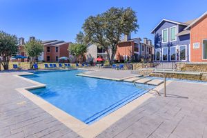 A sunny outdoor swimming pool area featuring a clear blue pool with lounge chairs, surrounded by greenery. In the background, there are several residential buildings with colorful exteriors, and shaded seating options under red umbrellas. The scene conveys a relaxing and inviting atmosphere.