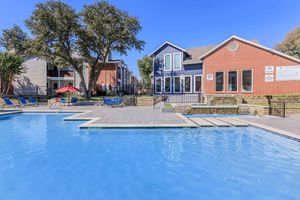 A bright blue swimming pool with lounge chairs surrounding it, in front of a modern building with a mix of brick and siding. The area is sunny, with trees nearby, and neighboring apartment buildings visible in the background. A shaded seating area with a red umbrella adds to the inviting atmosphere.