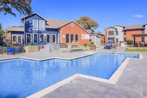 A clean, bright swimming pool with a surrounding patio area featuring blue lounge chairs. In the background, two colorful apartment buildings with a well-maintained landscape under a clear blue sky.