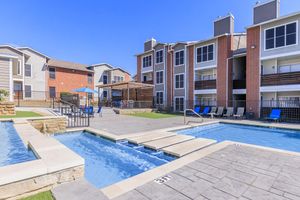 A well-designed outdoor pool area featuring two pools, one with a spa-like section. Surrounding the pools are lounge chairs and tables, with two apartment buildings in the background. The sky is clear and blue, creating a warm and inviting atmosphere.