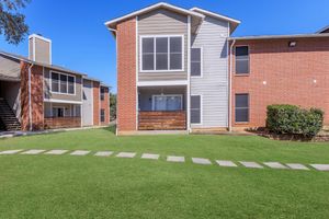 A view of an apartment complex featuring two buildings. One building has a modern grey and brick exterior with a balcony, while the other is predominantly brick. Well-kept green lawns and a stone pathway connect the two buildings under a clear blue sky.