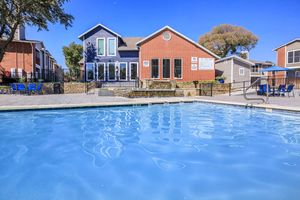 A calm swimming pool in the foreground reflecting the clear blue sky, with a modern, multi-colored building in the background. The building features large windows and outdoor seating areas. Surrounding the pool are lounge chairs and tables, creating a relaxing outdoor space.