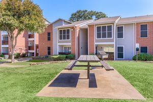 A landscaped courtyard featuring a picnic table on a concrete pad, surrounded by green grass and apartment buildings with screened porches. The buildings exhibit a mix of brick and siding exteriors, with trees providing shade in the area. The scene is bright and inviting under clear blue skies.
