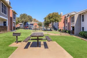 A landscaped courtyard featuring a picnic table on a concrete slab, surrounded by green grass. On either side, there are residential buildings, with some trees and a barbecue grill nearby. The sky is clear, creating a bright and inviting atmosphere.