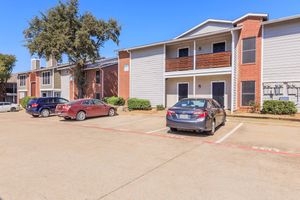 A view of a residential apartment complex featuring two buildings, parked cars in the foreground, and a clear blue sky. The buildings have a mix of brick and siding exteriors, with balconies visible on the upper levels. There are air conditioning units outside and landscaped areas around the parking lot.