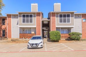 Two-story apartment building with a brick facade and light-colored siding. The entrance has large windows, and there is a white car parked in front. Green bushes flank the sides of the building, and parking spaces are marked in front. The scene is bright and sunny.