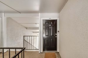 A view of an apartment building entrance featuring a black front door with a small window, surrounded by light-colored walls. A staircase to the left leads upward, and a welcome mat is placed in front of the door. The area is well-lit and clean, showcasing a typical residential setting.