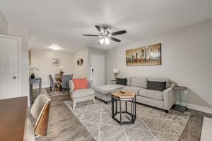 A cozy living room featuring a light gray sectional sofa with accent pillows, a wooden coffee table, and a decorative rug. There’s a ceiling fan, a small side table with a lamp, and a dining area in the background with a round table and chairs. The walls are decorated with modern art.
