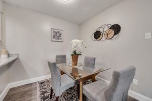 A modern dining area featuring a glass table surrounded by four gray upholstered chairs. A potted white orchid centerpiece sits on the table. The walls are a soft gray, adorned with a decorative wall hanging and a piece of artwork. A patterned rug adds warmth to the space.