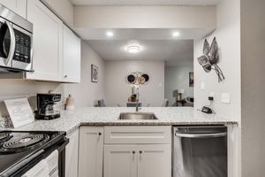 Modern kitchen featuring granite countertops, stainless steel appliances, and a coffee maker. There is a kitchen sink with a faucet, white cabinetry, and decorative wall art including leaf motifs. Soft lighting illuminates the space, creating a warm and inviting atmosphere.