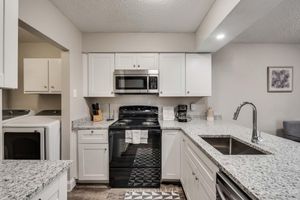Modern kitchen featuring white cabinetry, granite countertops, a stainless steel oven and microwave, and a black stove. A double sink is positioned in the island, which includes a dish rack in front. There’s a laundry area visible to the left, and a coffee maker on the counter. Decor includes a framed wall art piece.