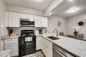 Modern kitchen with white cabinets, a black stove, and microwave. Countertops are covered in light gray speckled granite. Stainless steel sink with a sleek faucet, and a dining area in the background featuring a table with gray chairs and a decorative plant. Bright lighting enhances the space.