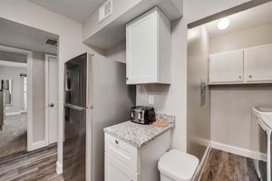 A modern kitchen interior featuring a stainless steel refrigerator, a toaster on a granite countertop, and white cabinetry. The space is well-lit, with a view into an adjoining room and laundry area visible in the background. The flooring is a dark laminate, contrasting with the light-colored walls.