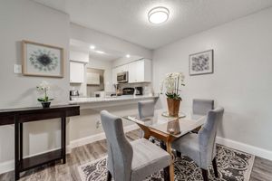 A modern dining area featuring a glass-top table surrounded by four gray upholstered chairs. There is a decorative plant with white flowers on the table, and wall art is visible in the background. The kitchen area is partially visible, showcasing sleek cabinetry and appliances.