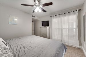 A cozy bedroom featuring a light gray wall, a queen-sized bed with a gray comforter, and a ceiling fan. There's a tall dresser against one wall, a flat-screen TV mounted on another, and large windows with sheer curtains allowing natural light to fill the room. A framed artwork is visible on the wall.